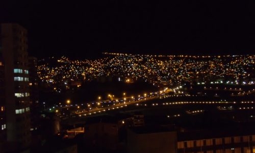 The city at night is lit up with lights on the buildings and street lights. The city is bustling with activity and the lights create a warm and inviting atmosphere. The buildings are tall