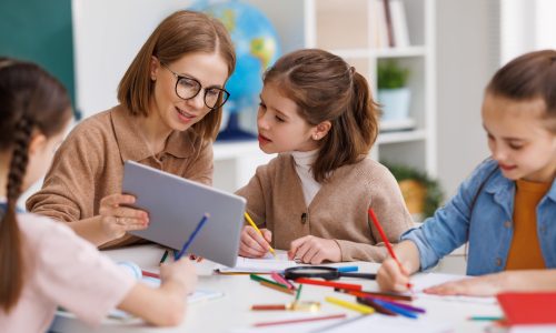 Happy female teacher in glasses smiling and looking at little student while sitting at table and showing data on tablet during lesson at school